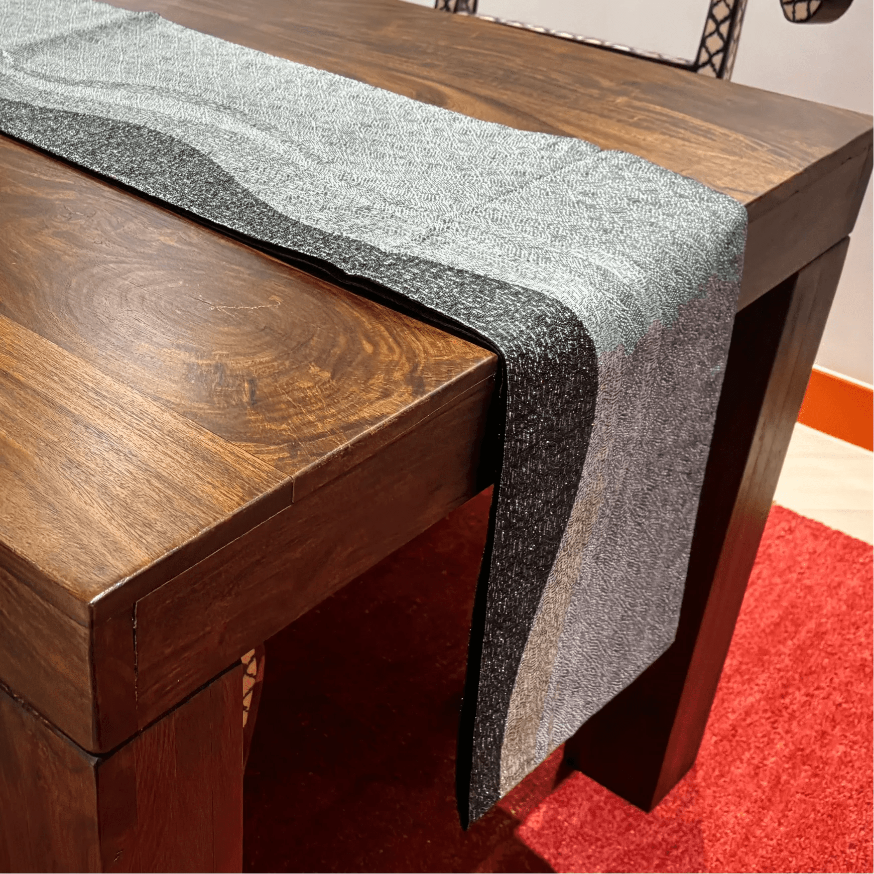 Wooden table with a silver and black patterned table runner on a red carpet.