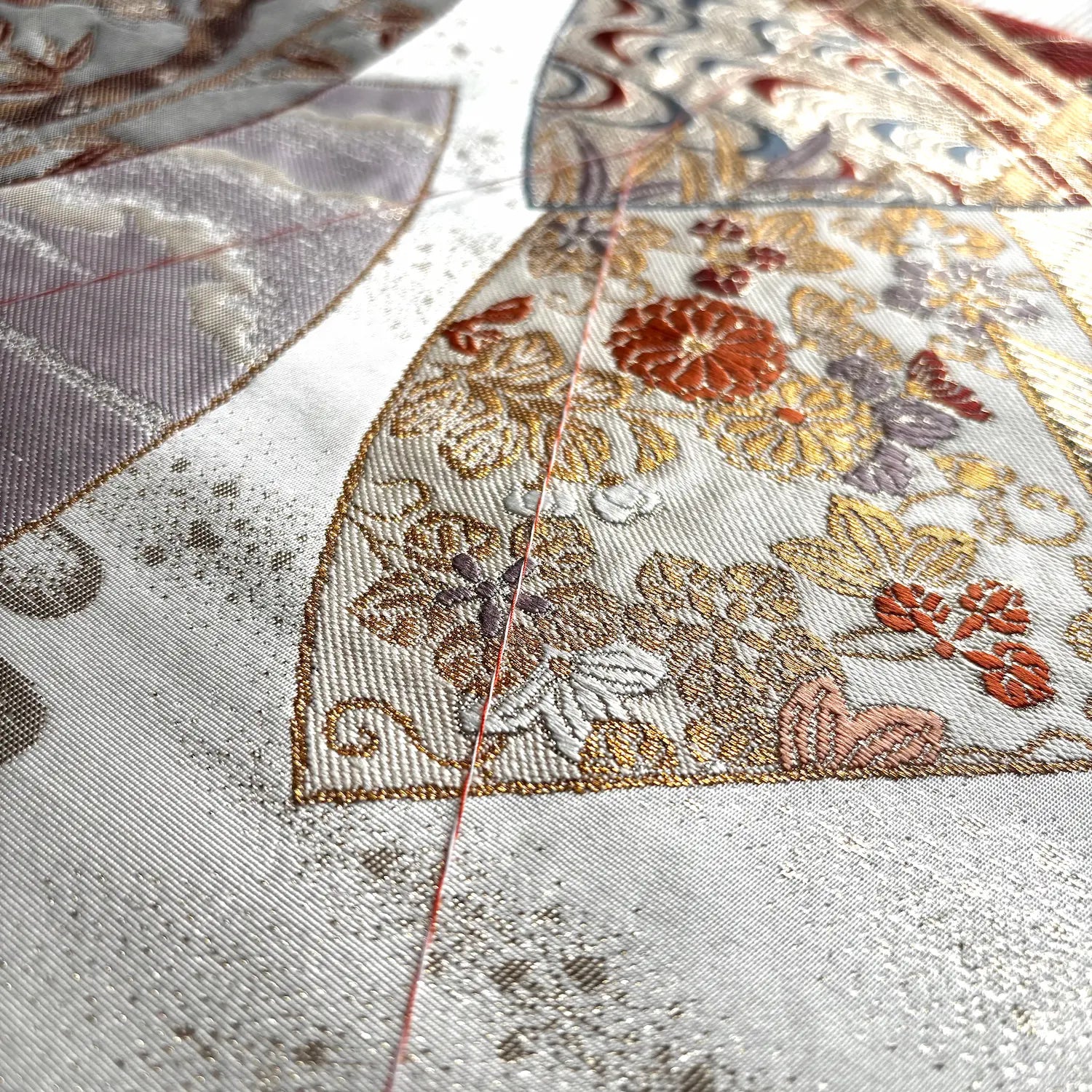 Close-up of an Kimono Obi with a cream base and detailed floral motifs in gold, rust red, taupe, and gray, showing fine woven texture and a thin red thread running vertically.