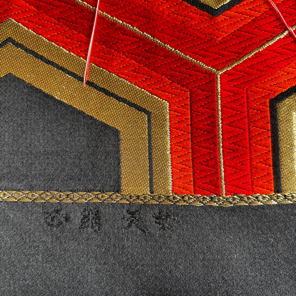 Close-up of a textured fabric product featuring a geometric pattern with red, gold, and black sections, finished with a braided gold trim and small embroidered characters on the black section.