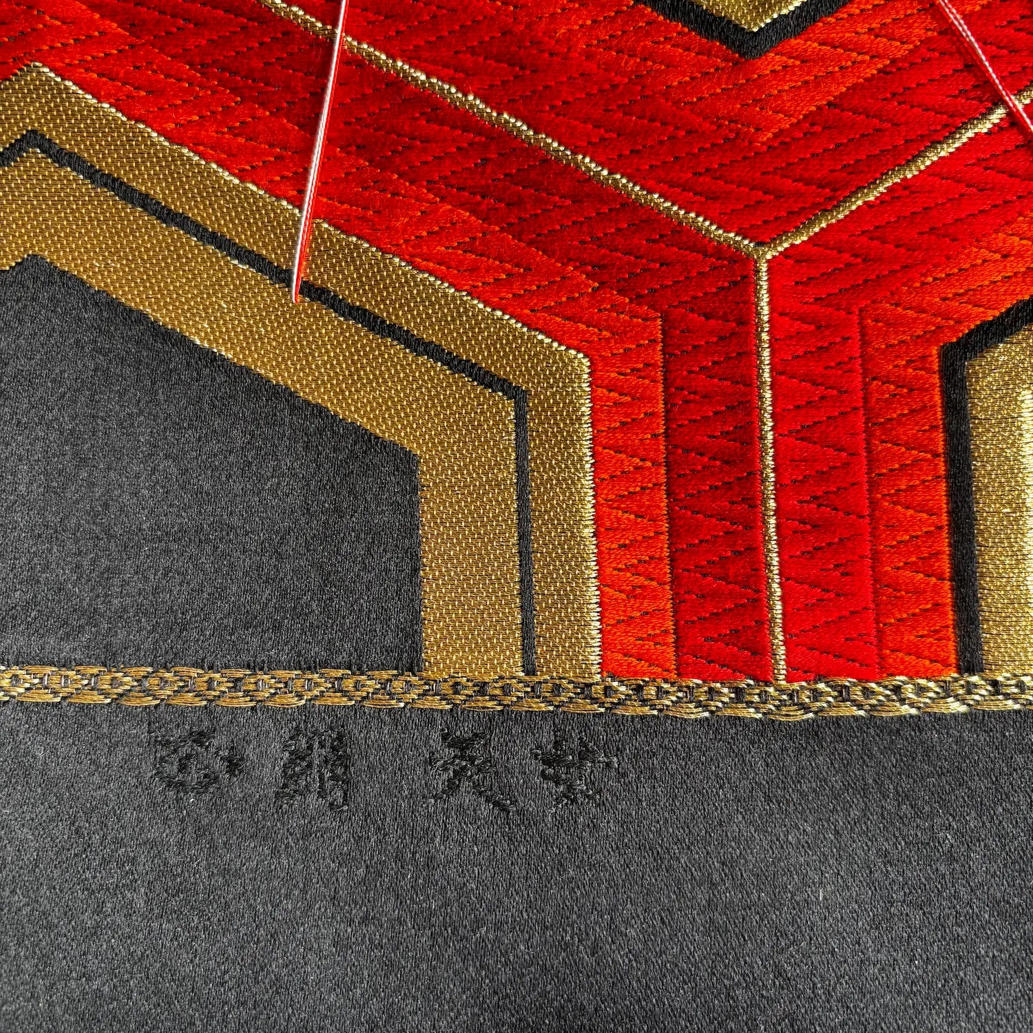 Close-up of a textured fabric product featuring a geometric pattern with red, gold, and black sections, finished with a braided gold trim and small embroidered characters on the black section.